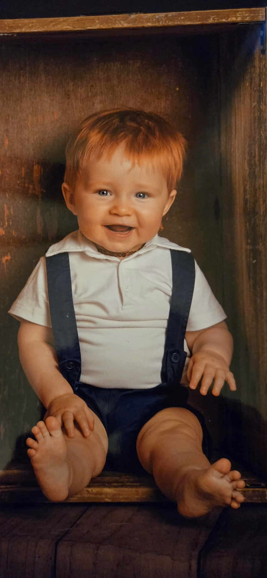 Maélyo a rejoint le concours — aidez-le/la à gagner de superbes lots ! baby, child, smiling, red_hair, blue_eyes, white_shirt, suspenders, wooden_box, feet, toes, happy, portrait, cute, infant, sitting, chubby_legs, indoors, closeup, hands, playful