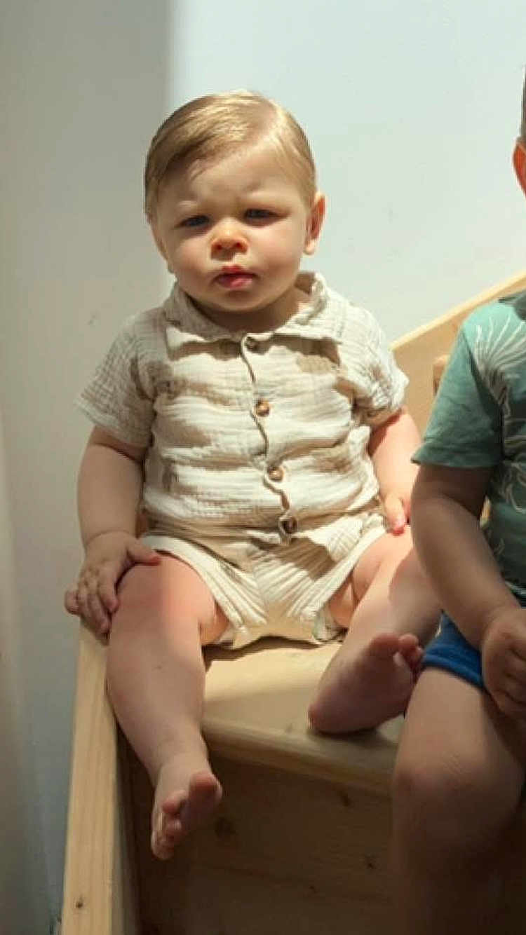 Ambrossio a rejoint le concours — aidez-le/la à gagner de superbes lots ! toddler, child, sitting, wooden_bench, beige_clothing, short_sleeves, shorts, indoor, natural_light, serious_expression, barefoot, person, face, skin, hair, light_shadows, furniture, two_children, casual_wear, young