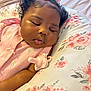 baby, sleeping, pink_clothing, bow, curly_hair, pacifier, pillow, floral_pattern, infant, peaceful, resting, cute, face, child, indoors, soft_lighting, closeup, skin, head, portrait