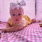 Raylin Honey is registered to the contest to win money with this photo: baby, headband, bow, yellow_outfit, pink_checkered_blanket, golden_eggs, infant, lying_down, curious_expression, blue_eyes, closeup, child, cute, soft_lighting, indoors, portrait, plush_background, small_hands, resting, decorative_eggs