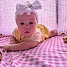 baby, headband, bow, yellow_outfit, pink_checkered_blanket, golden_eggs, infant, lying_down, curious_expression, blue_eyes, closeup, child, cute, soft_lighting, indoors, portrait, plush_background, small_hands, resting, decorative_eggs