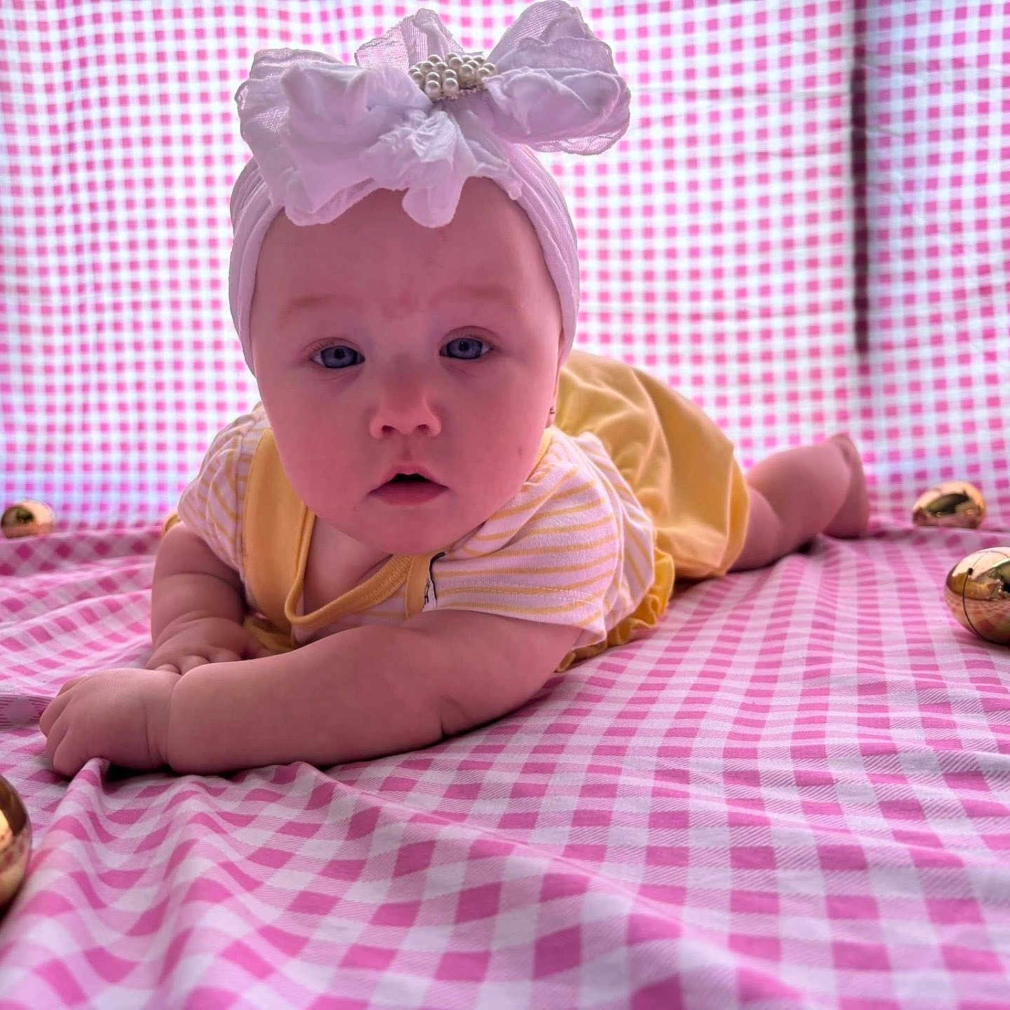 Raylin Honey is registered to the contest to win money with this photo: baby, headband, bow, yellow_outfit, pink_checkered_blanket, golden_eggs, infant, lying_down, curious_expression, blue_eyes, closeup, child, cute, soft_lighting, indoors, portrait, plush_background, small_hands, resting, decorative_eggs