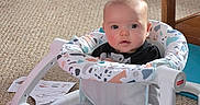 Cory is registered to the contest to win money with this photo: baby, baby_seat, baby_toys, carpet, carpeted_floor, child, clothing, curious, cute, face, footwear, furniture, home, indoor, infant, person, rosy_cheeks, sitting, toy, wooden_table