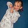 blue_background, brown_eyes, candid, child, curly_hair, ear, fingers, flash_photo, floral_dress, girl, hands, long_sleeves, playful, portrait, rosy_cheeks, smiling, studio_backdrop, teeth, white_dress, young_child