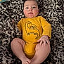 baby, infant, yellow_onesie, dinosaur_print, blanket, fuzzy_texture, leopard_print, child, cute, lying_down, soft_lighting, indoor, skin, face, legs, hand, foot, cozy, curious_expression, comfortable