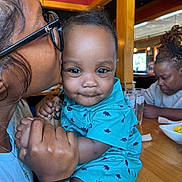 Jaleil is registered to the contest to win money with this photo: baby, person, kiss, glasses, dinosaur_print, turquoise_clothing, table, restaurant, wooden_table, hand, face, smile, indoor, adult, holding, people, background, female, casual, food