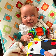 Noah joined the competition — help win amazing prizes! baby, smiling, tongue_out, toy, colorful, blanket, cushion, pattern, infant, happy, child, playful, hand, person, indoor, closeup, face, blue_eyes, white_clothing, soft_toys