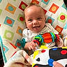 baby, smiling, tongue_out, toy, colorful, blanket, cushion, pattern, infant, happy, child, playful, hand, person, indoor, closeup, face, blue_eyes, white_clothing, soft_toys