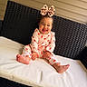 baby, child, infant, smiling, laughing, portrait, bow, hair_accessory, pajamas, pink_clothing, polka_dots, sitting, bed, cushion, outdoor_furniture, black_wicker, white_surface, happy, bare_feet, closeup