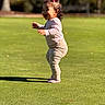 Liliana is registered to the contest to win money with this photo: toddler, child, baby, outdoor, park, grass, walking, smiling, pink_bow, hair, long_sleeves, pants, shoes, sunlight, shadow, lawn, portrait, profile, playful, daytime
