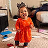 Liliana is registered to the contest to win money with this photo: toddler, child, baby, smiling, happy, standing, portrait, indoors, living_room, orange_dress, hair_bow, patterned_clothing, leggings, socks, carpet, laundry, toys, furniture, home, photography
