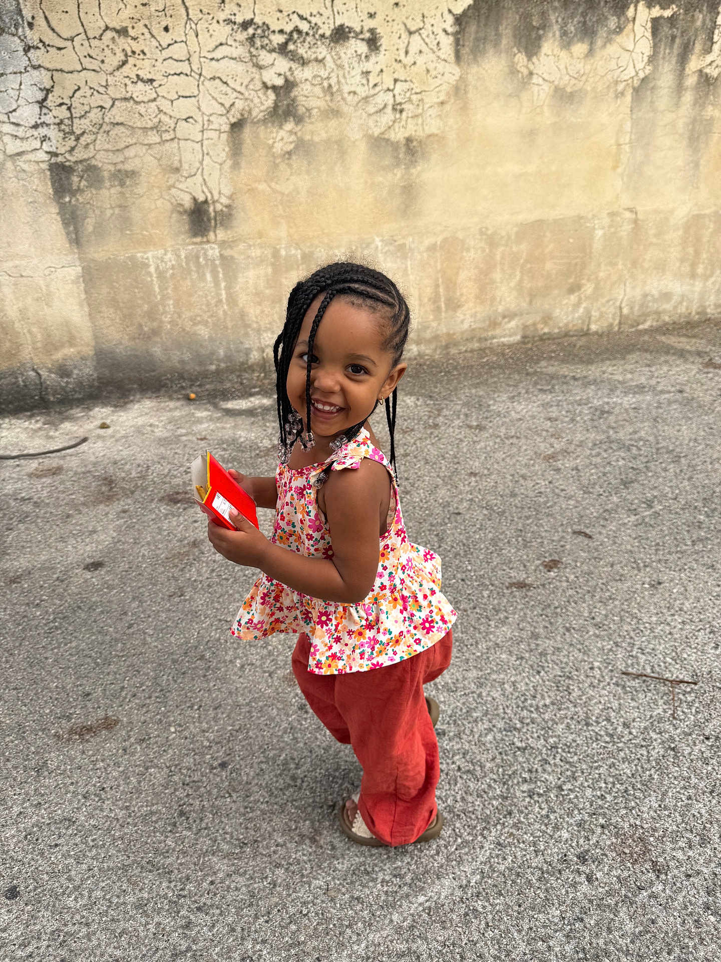 Daya a rejoint le concours — aidez-le/la à gagner de superbes lots ! child, smiling, braids, beads, floral_top, pants, sandals, outdoor, concrete, wall, joyful, cute, standing, playful, person, young, happy, fashion, portrait, daylight