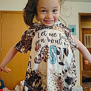 Jaylee is registered to the contest to win money with this photo: child, smiling, indoor, tshirt, animal_print, text, long_hair, messy_hair, playful, door, carpet, room, clothing, person, happy, home, casual, young, portrait, cute
