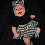 baby, infant, smiling, headband, bow, houndstooth_pattern, black_background, black_clothing, dress, skirt, booties, lying_down, happy, chubby_cheeks, fist, legs, portrait, closeup, eyes, mouth_open