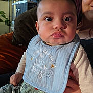 Elijah joined the competition — help win amazing prizes! baby, child, person, bib, drool, indoor, smile, clothing, face, hand, adult, couch, home, cute, expression, portrait, sitting, long_sleeve, pants, patterned_clothing