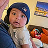 Elijah is registered to the contest to win money with this photo: baby, child, face, hat, baseball_cap, person, indoor, living_room, couch, cushions, painting, expression, cute, curious, holding, clothing, portrait, skin, eyelashes, hand