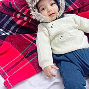 Rahid is registered to the contest to win money with this photo: baby, child, infant, fur_hood, knitted_sweater, bear_design, plaid_blanket, red_blanket, black_blanket, blue_pants, smiling, cute, cozy, warm_clothing, portrait, indoor, lying_down, hand, face, eyes