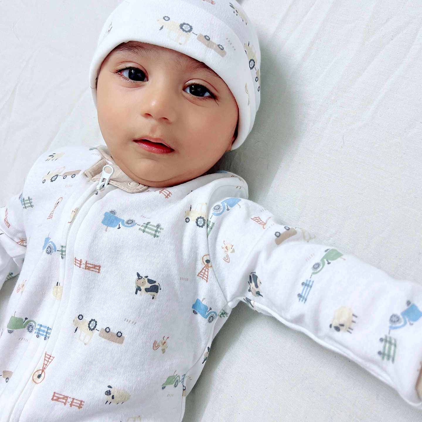 Rahid is registered to the contest to win money with this photo: animal_print, baby, bed_sheet, black_eyes, child, curious_expression, cute, face, farm_print, hat, indoors, infant, lying_down, onesie, portrait, sleepwear, soft_lighting, tractor_print, white_clothing, young_child