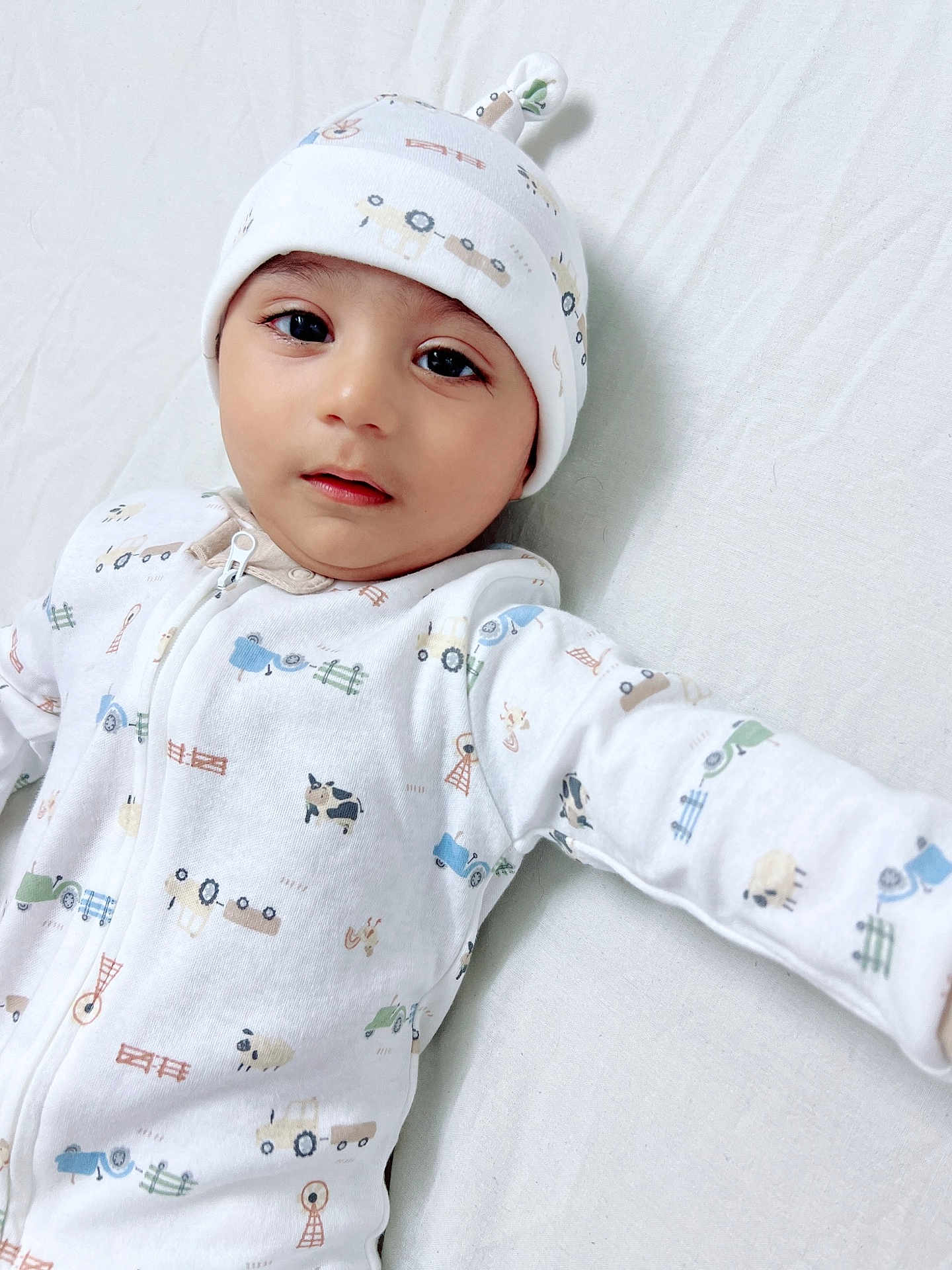 Rahid is registered to the contest to win money with this photo: baby, infant, onesie, hat, white_clothing, farm_print, tractor_print, animal_print, lying_down, bed_sheet, child, cute, face, black_eyes, sleepwear, soft_lighting, portrait, young_child, indoors, curious_expression