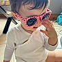 toddler, child, sunglasses, pink, heart_pattern, white_clothing, indoor, floor, hand, hair, cute, playing, object, natural_light, casual, baby, sitting, person, portrait, expression