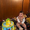 baby, birthday, birthday_cake, cake, child, colorful, celebration, dessert, frosting, food, indoors, infant, party, portrait, seated, smiling, table, toy_character, wood_paneling, cake_decorations