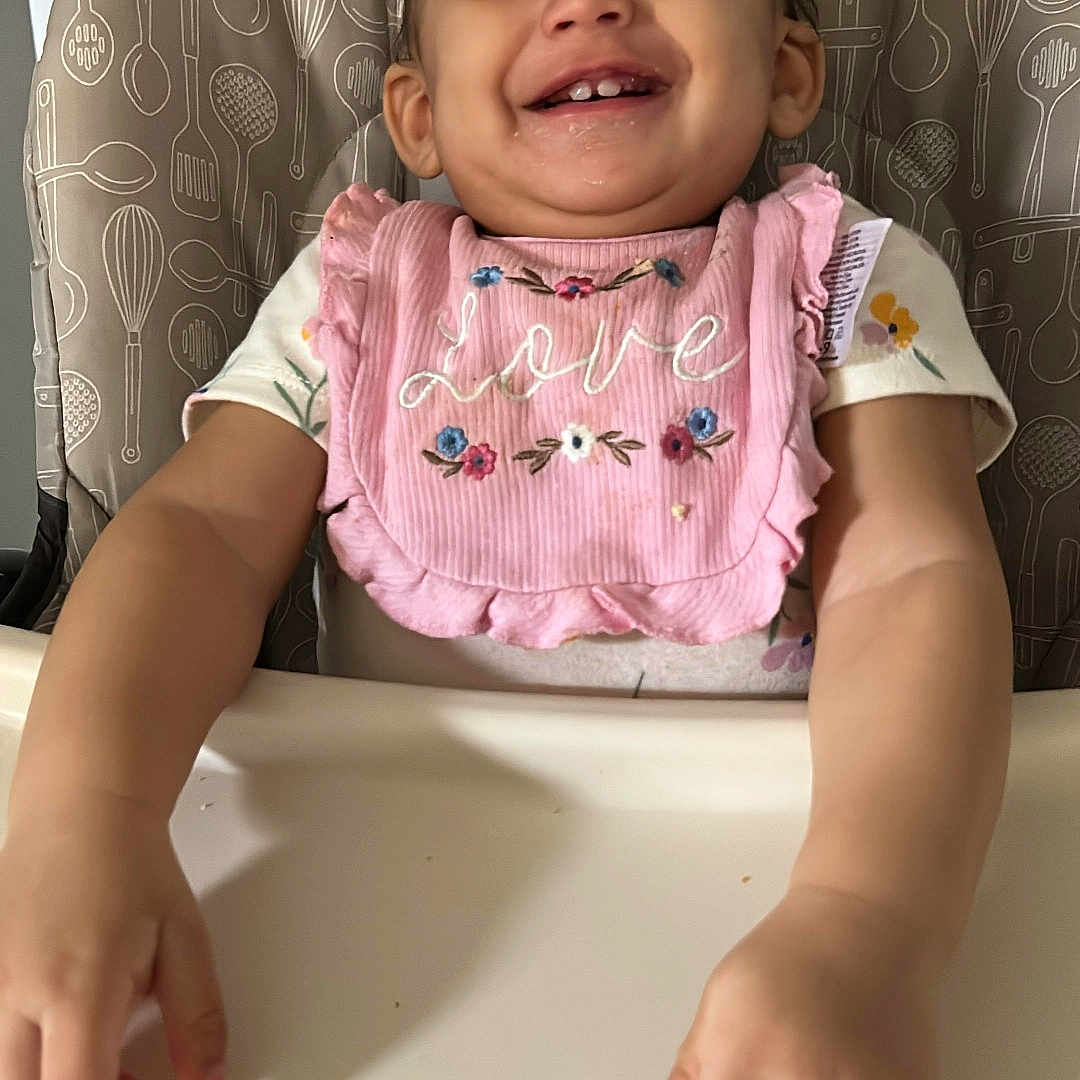 Jocabed is registered to the contest to win money with this photo: baby, bib, child, clothing, cute, expression, face, feeding, food, hands, happy, high_chair, indoor, meal, person, pink, smiling, snack, table, toddler
