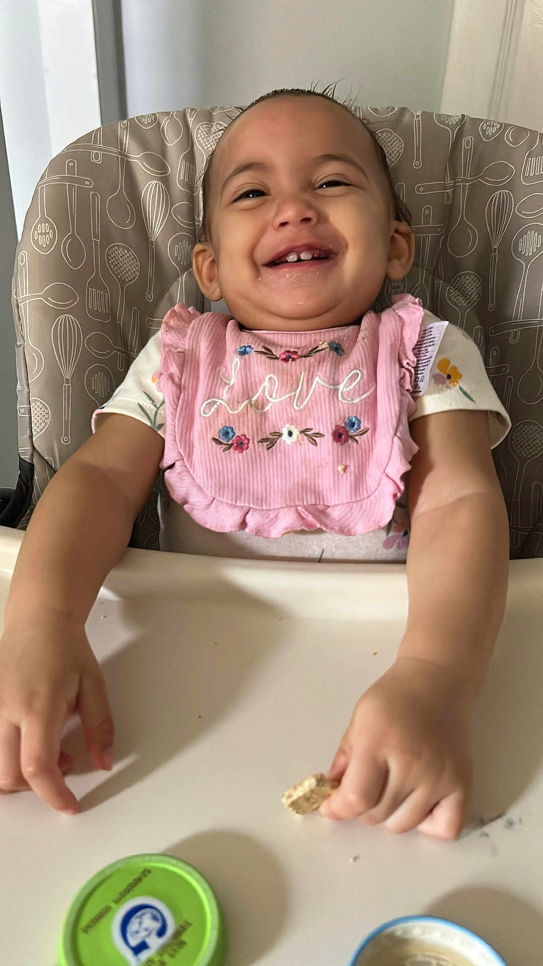 Jocabed is registered to the contest to win money with this photo: toddler, child, high_chair, bib, pink, smiling, food, snack, happy, cute, baby, face, hands, clothing, indoor, person, table, meal, expression, feeding