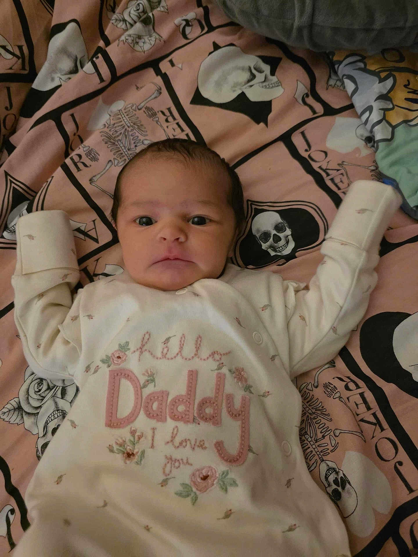 Seren is registered to the contest to win money with this photo: baby, infant, onesie, clothing, blanket, skull_pattern, skeleton_pattern, face, person, lying_down, cute, embroidery, floral_design, text_on_clothing, bedroom, indoors, newborn, expression, wide_eyes, curious