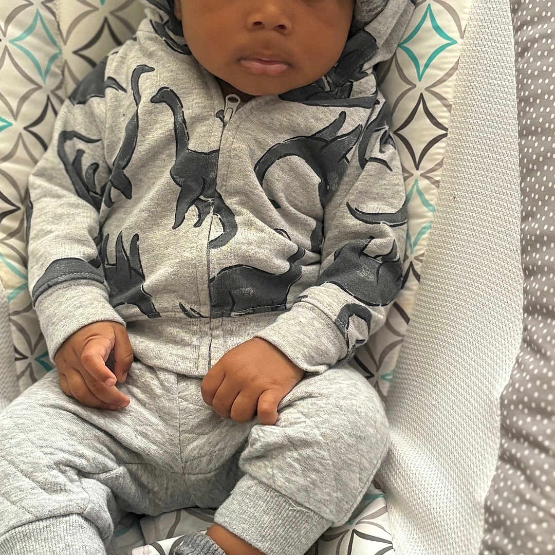 Ali is registered to the contest to win money with this photo: baby, casual_wear, child, comfortable, cozy, cute, dinosaur_print, gray_clothing, hat, hoodie, indoors, infant, knit_hat, looking_at_camera, patterned_seat, portrait, sitting, socks, toddler, young_child