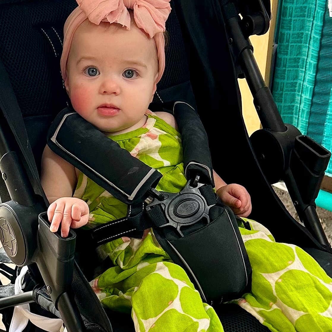 Mia joined the competition — help win amazing prizes! baby, child, stroller, headband, pink_bow, green_outfit, barefoot, infant, seatbelt, curious_look, indoor, casual_clothing, foot, face, person, cute, young_child, sitting, portrait, baby_clothes