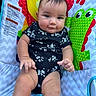 baby, infant, child, onesie, black_clothing, floral_pattern, play_mat, colorful, cartoon_animal, frog, yellow_bird, cheeks, expression, sitting, hand, legs, skin, indoor, portrait, cute