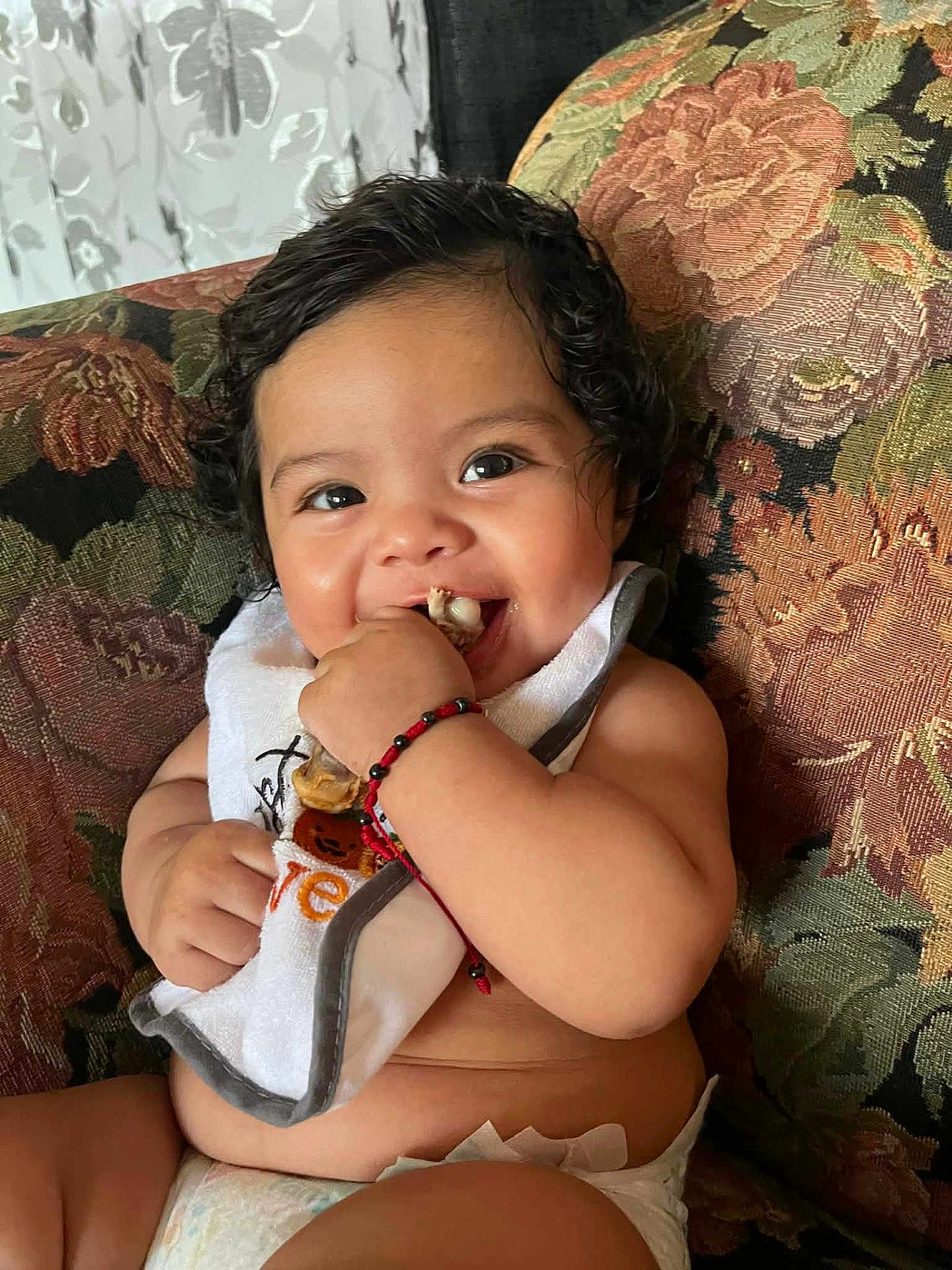 Adamaris is registered to the contest to win money with this photo: baby, child, smiling, curly_hair, bib, diaper, sitting, toy, happy, indoor, couch, floral_pattern, cute, cheerful, skin, hand, face, infant, playful, young