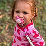 toddler, child, pacifier, red_hair, pink_shirt, hearts, outdoor, greenery, smiling, happy, cute, beads, clothing, person, portrait, baby, young_child, playful, nature, closeup
