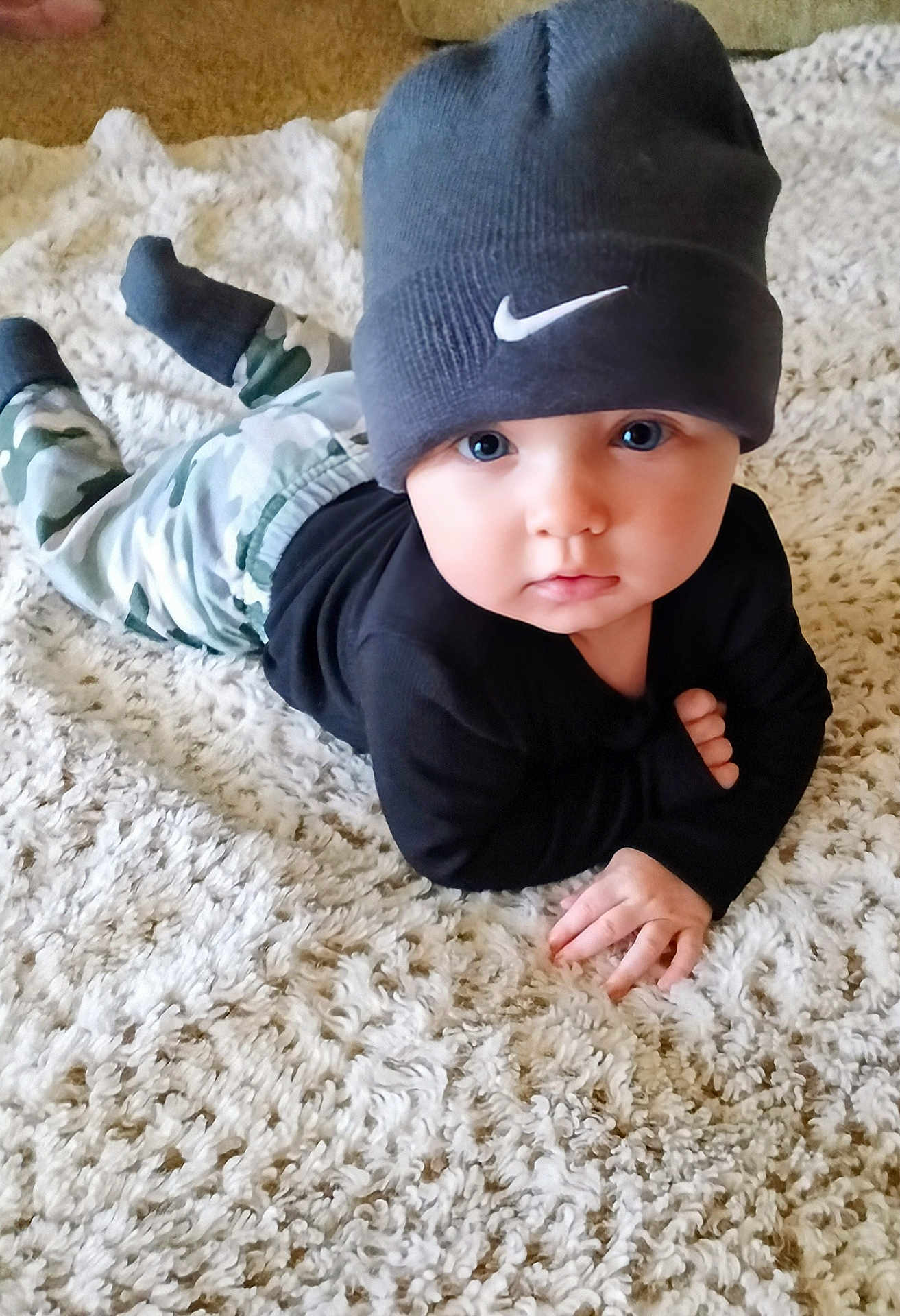 Kane is registered to the contest to win money with this photo: baby, child, beanie, hat, carpet, camo_pants, black_top, lying_down, indoors, curious, blue_eyes, cute, soft_texture, hand, face, portrait, infant, fashion, young_child, floor