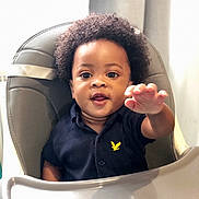 Jason is registered to the contest to win money with this photo: toddler, child, curly_hair, high_chair, navy_shirt, yellow_emblem, reaching_hand, indoor, soft_light, cute, happy, baby, portrait, person, seat, expression, young_child, smiling, skin, face