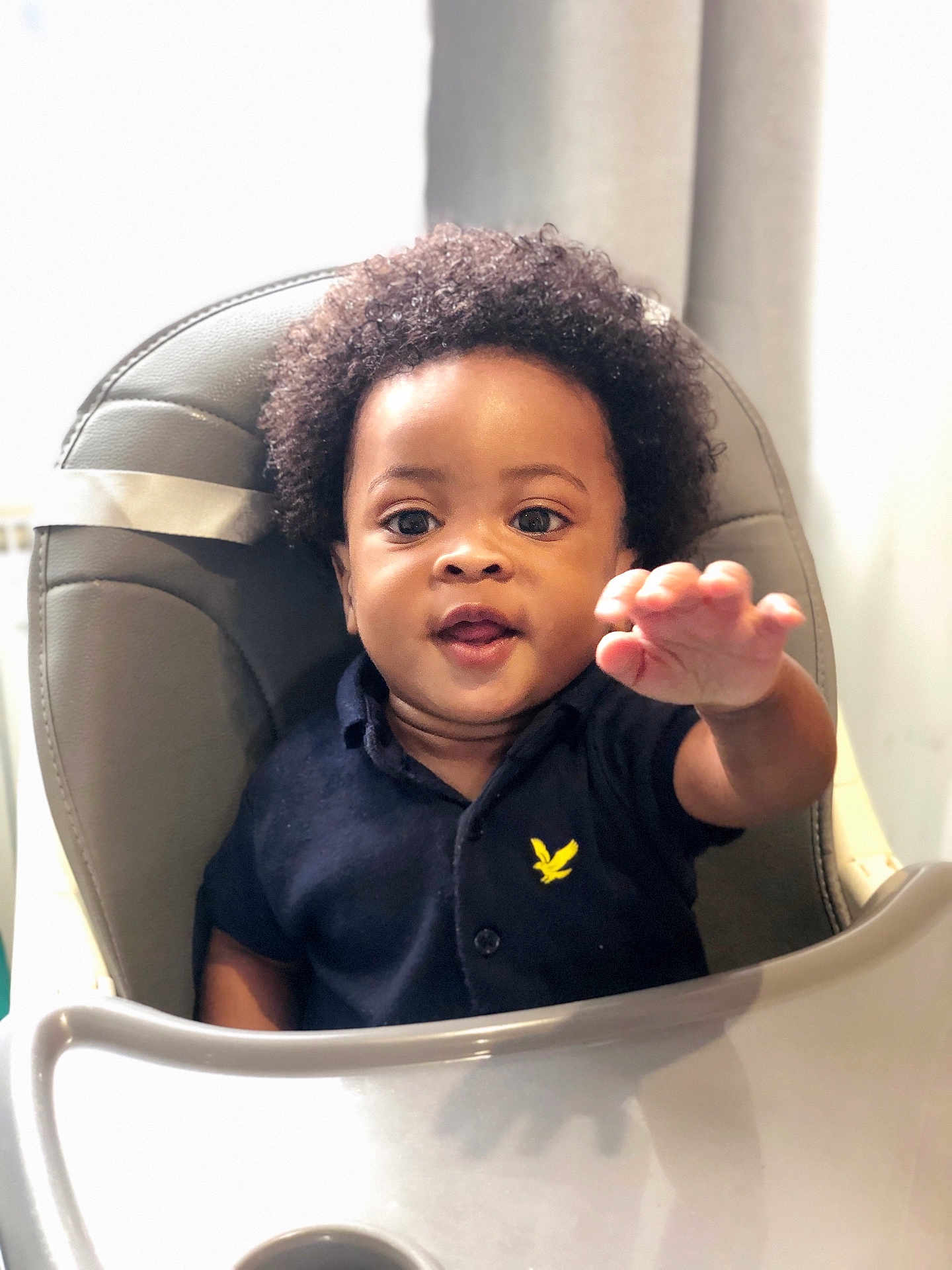 Jason is registered to the contest to win money with this photo: toddler, child, curly_hair, high_chair, navy_shirt, yellow_emblem, reaching_hand, indoor, soft_light, cute, happy, baby, portrait, person, seat, expression, young_child, smiling, skin, face