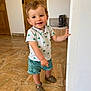 toddler, child, smiling, indoor, standing, shirt, shorts, sandals, floor, door, hand, face, person, curly_hair, expression, young, home, marble_floor, wooden_door, playful
