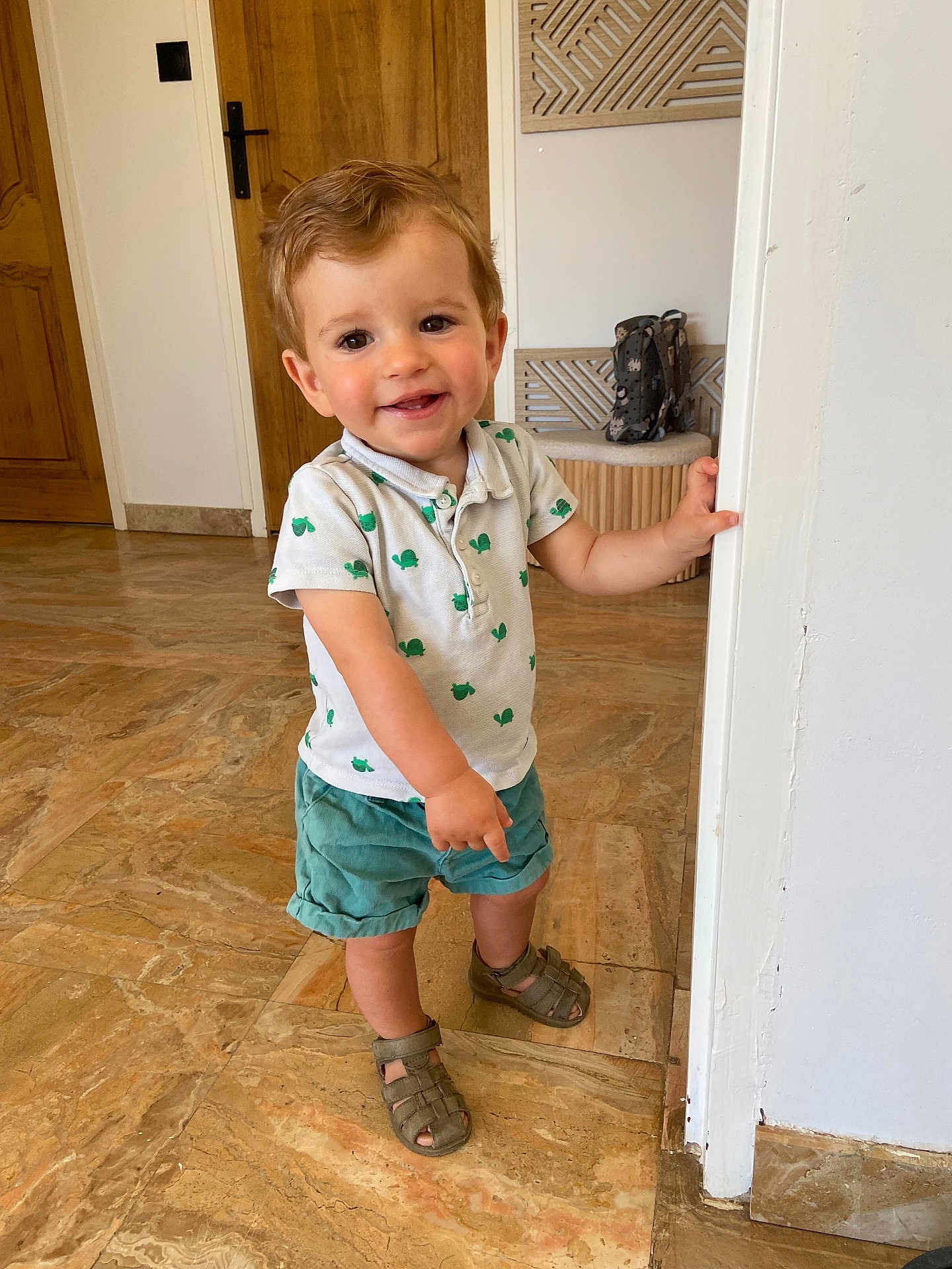 Raphaël participe au concours pour gagner de l'argent avec cette photo : toddler, child, smiling, indoor, standing, shirt, shorts, sandals, floor, door, hand, face, person, curly_hair, expression, young, home, marble_floor, wooden_door, playful