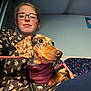 person, dog, glasses, indoor, leash, harness, seated, floral_clothing, spotted_dog, pet, casual, public_transport, blue_seat, sign, curious, closeup, warm_lighting, candid, portrait, young_adult