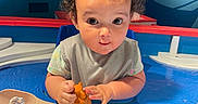 Skyla joined the competition — help win amazing prizes! toddler, child, water_play, toy, orange_toy, bow, curly_hair, blue_background, water_fountain, play_area, indoor, water, sitting, cute, face, expression, short_sleeves, bib, hands, curious