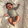 Skyla is registered to the contest to win money with this photo: baby, infant, child, onesie, gray_clothing, bed, white_bedding, pillow, smiling, happy, lying_down, indoors, cute, playful, person, skin, face, hair, feet, hands