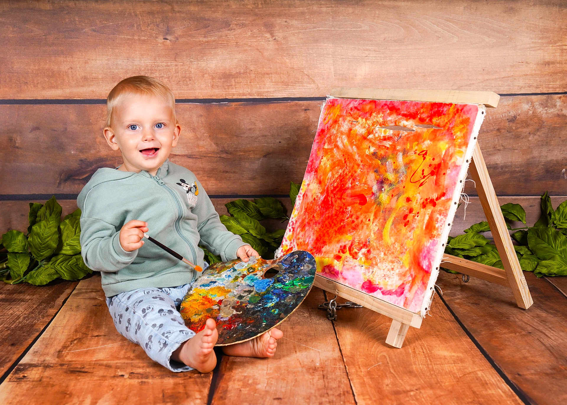 Kaysan participe au concours pour gagner de l'argent avec cette photo : toddler, child, painting, palette, paintbrush, artwork, easel, wooden_floor, wooden_background, green_leaves, smiling, barefoot, sitting, hoodie, pants, indoor, colorful, creative, portrait, happy