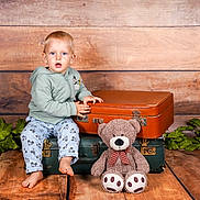 Kaysan a rejoint le concours — aidez-le/la à gagner de superbes lots ! toddler, child, baby, suitcase, vintage_suitcase, teddy_bear, plush_toy, wooden_floor, wooden_wall, green_leaves, pants, hoodie, barefoot, indoor, portrait, seated, curious, clothing, toy, photography