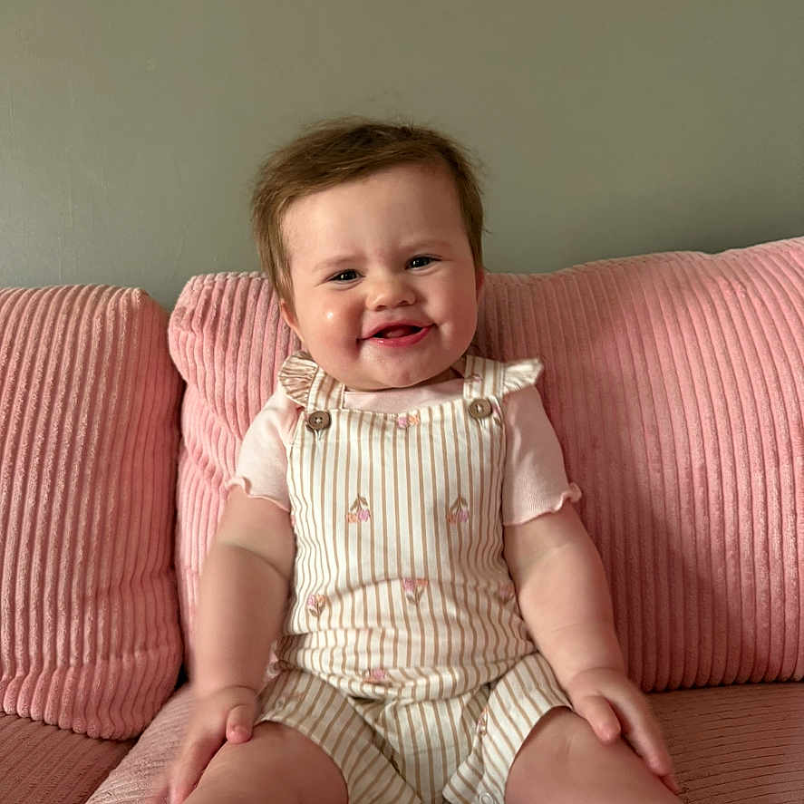 Layken joined the competition — help win amazing prizes! baby, child, smiling, couch, pink, pillow, overalls, shorts, striped, indoor, person, happy, seated, furniture, cute, portrait, infant, clothing, home, relaxed