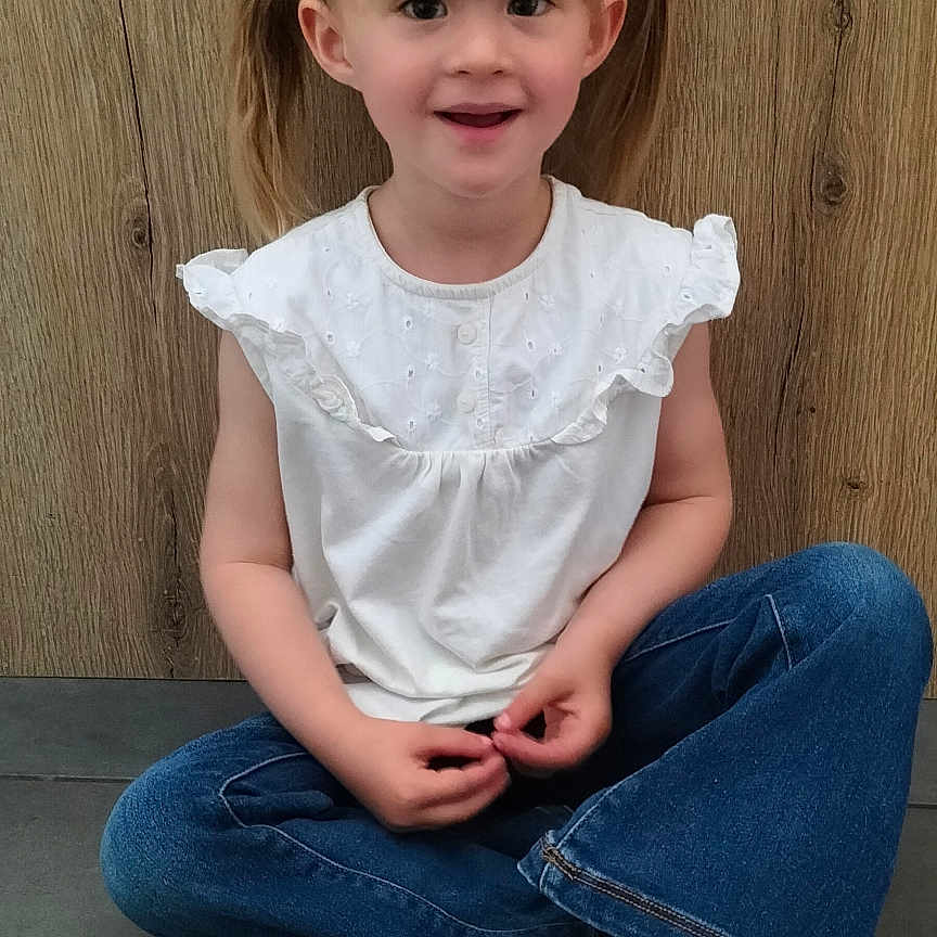 Giulia a rejoint le concours — aidez-le/la à gagner de superbes lots ! blonde_hair, blue_jeans, blue_shoes, casual_clothing, child, cross_legged, cute, floor_tiles, floral_bow, girl, happy, indoor, person, pigtails, portrait, sitting, smiling, white_top, wooden_wall, young