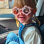 child, girl, sunglasses, flower_shaped_glasses, braided_hair, denim_overalls, white_shirt, car_interior, seat, sunlight, smile, happy, portrait, casual_clothing, reflection, window, seatbelt, young_child, indoor, daylight