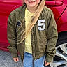 child, girl, red_headband, blonde_hair, green_jacket, yellow_top, blue_jeans, white_shoes, fluffy_slippers, car, red_car, car_wheel, outdoor, sidewalk, smiling, portrait, casual_clothing, daylight, standing, happy