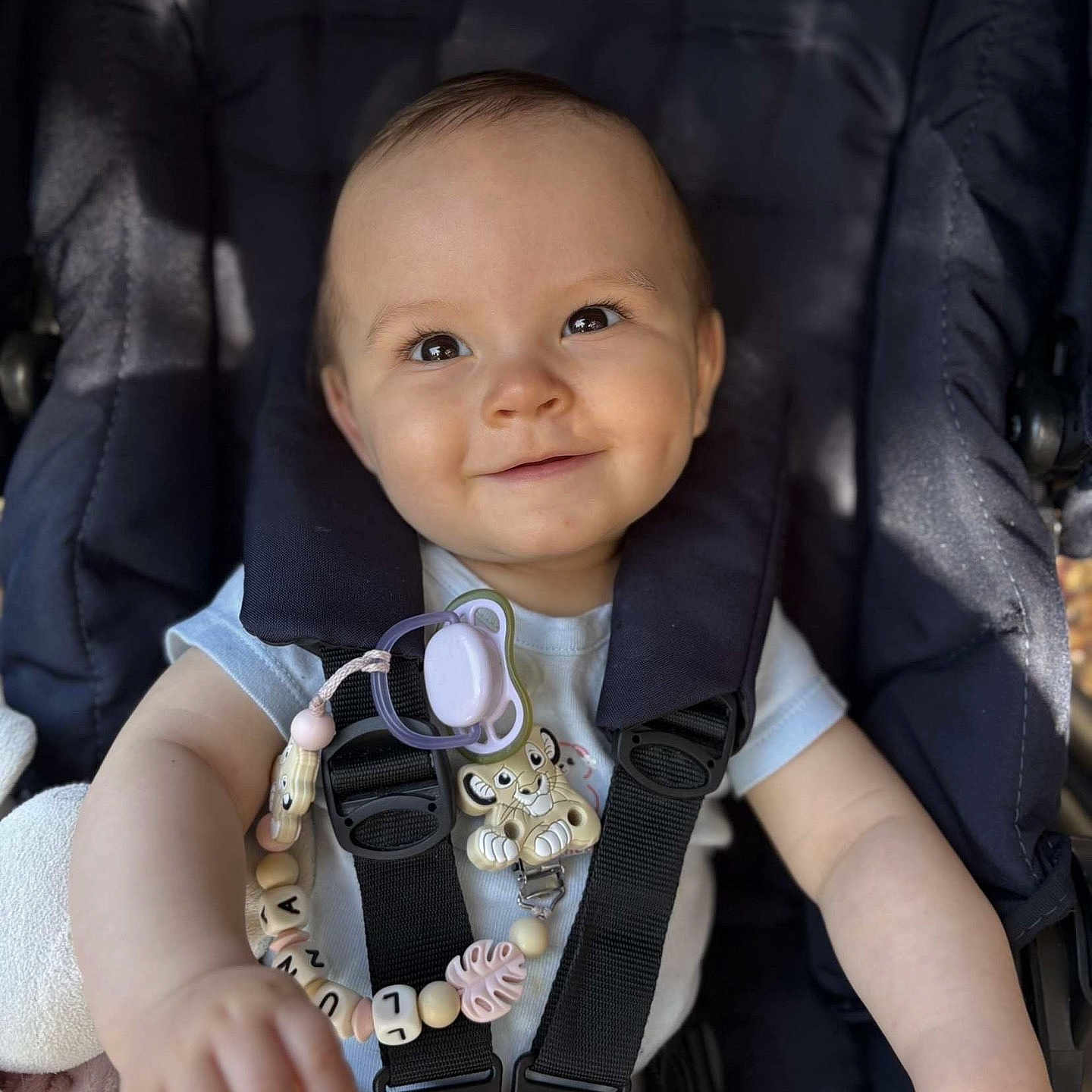 Luna participe au concours pour gagner de l'argent avec cette photo : baby, baby_accessory, black_straps, child, clip, comfort, cute, dimples, face, hand, happy, infant, light_blue_shirt, lion, pacifier, portrait, safety_harness, seat, smiling, stroller