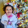 Mariah is registered to the contest to win money with this photo: child, smiling, doctor_costume, toy, stethoscope, christmas_tree, holiday_lights, decorations, indoor, carpet, festive, playful, young_child, curly_hair, white_coat, blue_slippers, red_bow, ornaments, holiday_season, joyful