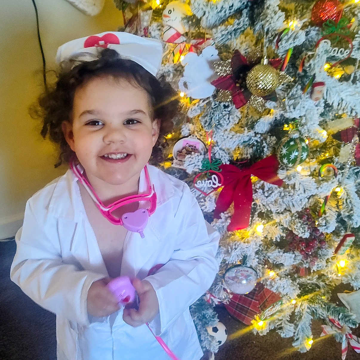 Mariah is registered to the contest to win money with this photo: child, smiling, doctor_costume, toy, stethoscope, christmas_tree, holiday_lights, decorations, indoor, carpet, festive, playful, young_child, curly_hair, white_coat, blue_slippers, red_bow, ornaments, holiday_season, joyful